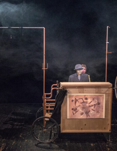 zdjęcie ze spektaklu teatru rawa momo na scenie wóz teatralny ze skrzynią na której podświetlona jest ilustracja za wozem ewa kubiak gra z lalką szarego pana